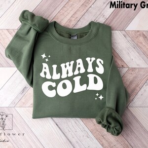 May include: A green sweatshirt with the text "ALWAYS COLD" in white retro-style lettering. The sweatshirt has a crew neck and long sleeves.