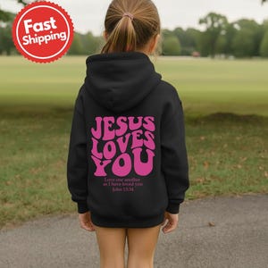 May include: A black hooded sweatshirt with the text "JESUS LOVES YOU" in pink, with the Bible verse John 13:34 below. The sweatshirt is on a child, with a green field in the background.