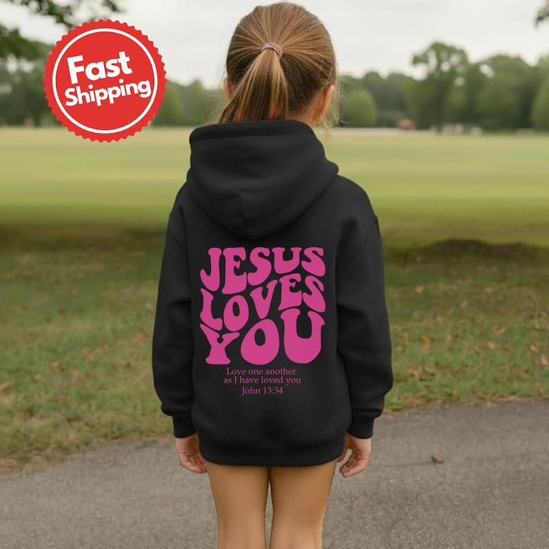 May include: A black hooded sweatshirt with the text "JESUS LOVES YOU" in pink, with the Bible verse John 13:34 below. The sweatshirt is on a child, with a green field in the background.