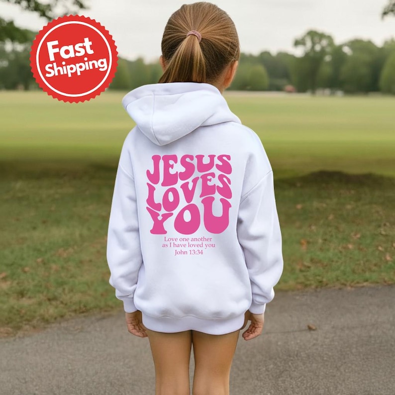 May include: A white hooded sweatshirt with the text "JESUS LOVES YOU" in pink, with the Bible verse John 13:34 below. The sweatshirt is on a person standing outside on a green lawn.