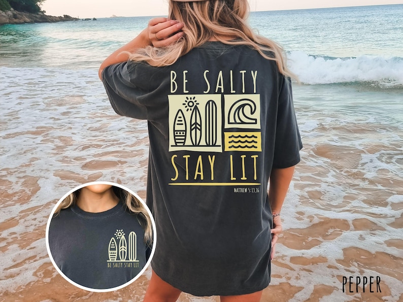 May include: A black t-shirt with a graphic design of surfboards, a sun, and waves. The text "BE SALTY STAY LIT" is printed on the shirt. The shirt is being worn by a person standing on a beach.