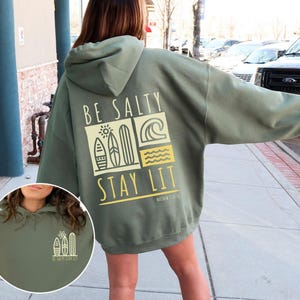 Może przedstawiać: Zielona bluza z kapturem z napisem "Be Salty Stay Lit" i grafiką desek surfingowych i fal na plecach. Bluza jest oversize'owa i ma luźny kr&oacute;j.