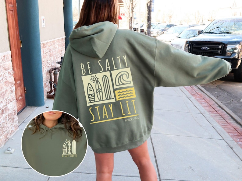 Może przedstawiać: Zielona bluza z kapturem z napisem "Be Salty Stay Lit" i grafiką desek surfingowych i fal na plecach. Bluza jest oversize'owa i ma luźny kr&oacute;j.