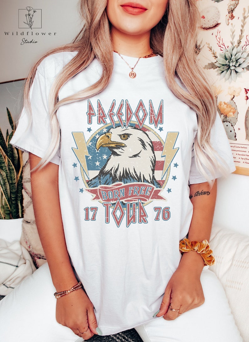Puede incluir: Camiseta blanca con un gr&aacute;fico de estilo vintage de un &aacute;guila con las palabras "Freedom Born Free 17 Tour 76" y una bandera estadounidense desgastada.