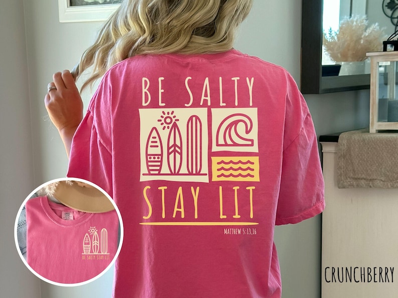 May include: Pink short-sleeved t-shirt with a graphic design that says "Be Salty Stay Lit" with a sun, waves, and surfboards. The text "Matthew 5:13-14" is printed below the graphic.