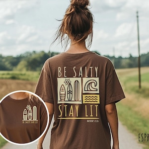 Może przedstawiać: Brązowa koszulka z graficznym wzorem desek surfingowych i tekstem "BE SALTY STAY LIT" w kolorze ż&oacute;łtym. Wz&oacute;r zawiera r&oacute;wnież tekst "MATTHEW 5:13".