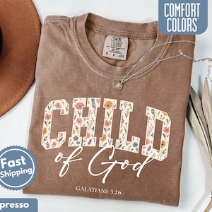 May include: Espresso-colored t-shirt with the words "CHILD of God" in a floral design. The text "GALATIANS 3:26" is below the main text. The shirt has a Comfort Colors tag and a Fast Shipping badge.