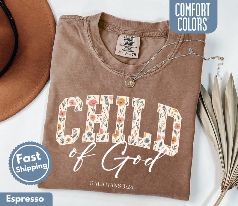 May include: Espresso-colored t-shirt with the words "CHILD of God" in a floral design. The text "GALATIANS 3:26" is below the main text. The shirt has a Comfort Colors tag and a Fast Shipping badge.
