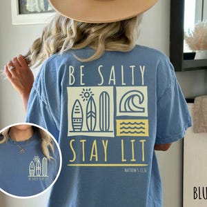 Może przedstawiać: Koszulka z niebieskiego dżinsu z napisem "Be Salty Stay Lit" i grafiką przedstawiającą deski surfingowe i fale. Na koszulce znajduje się r&oacute;wnież napis "Matthew 5:13-16".
