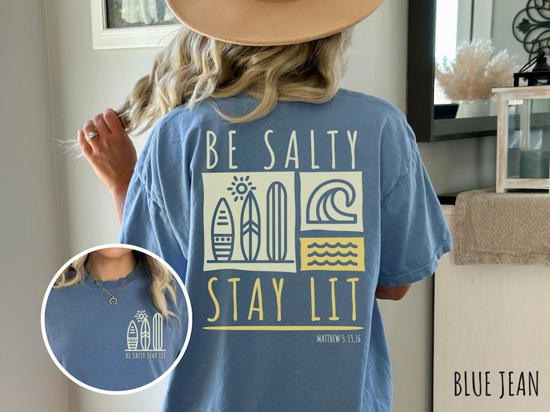 Może przedstawiać: Koszulka z niebieskiego dżinsu z napisem "Be Salty Stay Lit" i grafiką przedstawiającą deski surfingowe i fale. Na koszulce znajduje się r&oacute;wnież napis "Matthew 5:13-16".
