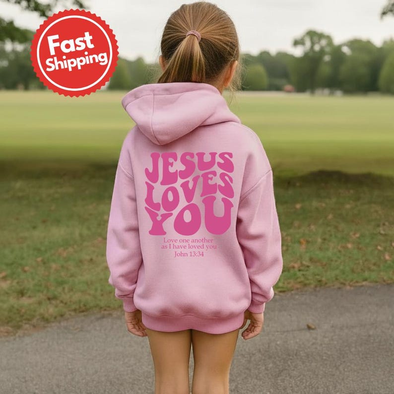 May include: A light pink hoodie with the text "JESUS LOVES YOU" in a retro font on the back. The hoodie also has the text "Love one another as I have loved you John 13:34".