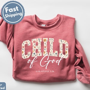 May include: Heather scarlet red sweatshirt with the words "CHILD of God" in a floral design. The text "Galatians 3:26" is below the main text. A blue and white "Fast Shipping" sticker is in the upper left corner.