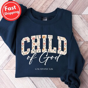 May include: Navy blue sweatshirt with the words "CHILD of God" in a floral design. The text "GALATIANS 3:26" is below the main text. A red and white "Fast Shipping" sticker is in the upper left corner.