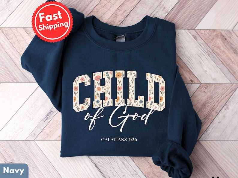 May include: Navy blue sweatshirt with the words "CHILD of God" in a floral design. The text "GALATIANS 3:26" is below the main text. A red and white "Fast Shipping" sticker is in the upper left corner.