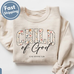 May include: Sand-colored sweatshirt with the words "CHILD of God" in a floral design. The text "GALATIANS 3:26" is below the main text. The sweatshirt has long sleeves and a crew neck.