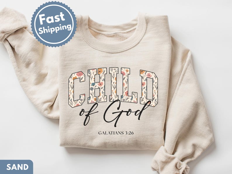May include: Sand-colored sweatshirt with the words "CHILD of God" in a floral design. The text "GALATIANS 3:26" is below the main text. The sweatshirt has long sleeves and a crew neck.