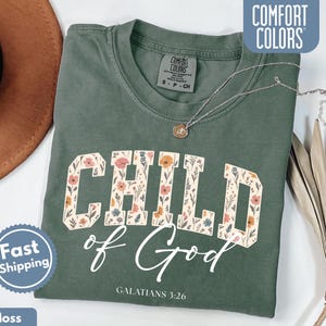 May include: A moss green t-shirt with the words "CHILD of God" in a floral design. The text "GALATIANS 3:26" is below the main text. A silver necklace with a pendant is on the shirt. The shirt is labeled "COMFORT COLORS".