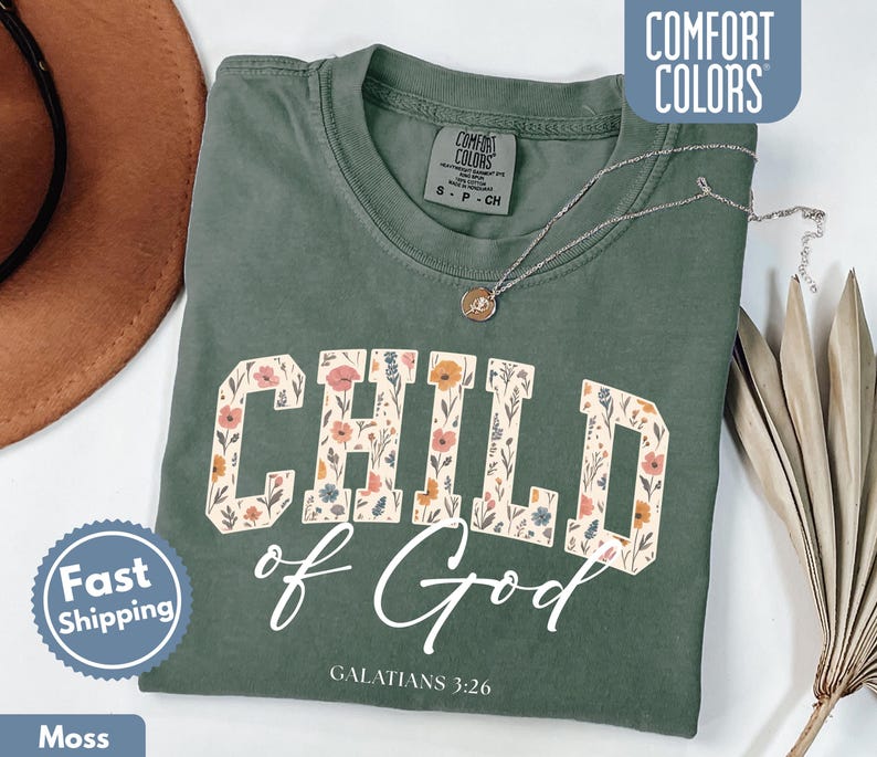 May include: A moss green t-shirt with the words "CHILD of God" in a floral design. The text "GALATIANS 3:26" is below the main text. A silver necklace with a pendant is on the shirt. The shirt is labeled "COMFORT COLORS".