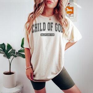 May include: A cream colored oversized t-shirt with the text "CHILD OF GOD" in black lettering and the text "GALATIANS 3:26" in smaller black lettering. The shirt is being worn by a person with long blonde hair.