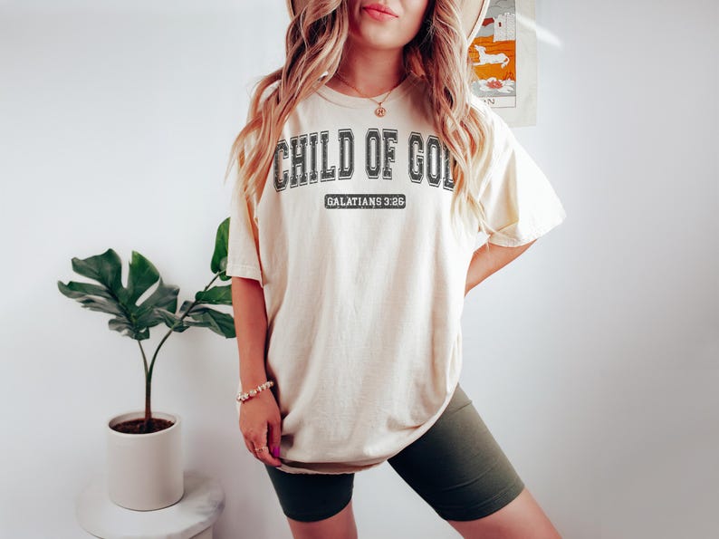 May include: A cream colored oversized t-shirt with the text "CHILD OF GOD" in black lettering and the text "GALATIANS 3:26" in smaller black lettering. The shirt is being worn by a person with long blonde hair.