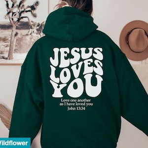 May include: A dark green hoodie with the text "JESUS LOVES YOU" in white, retro-style lettering on the back. Below the text is the quote "Love one another as I have loved you John 13:34".