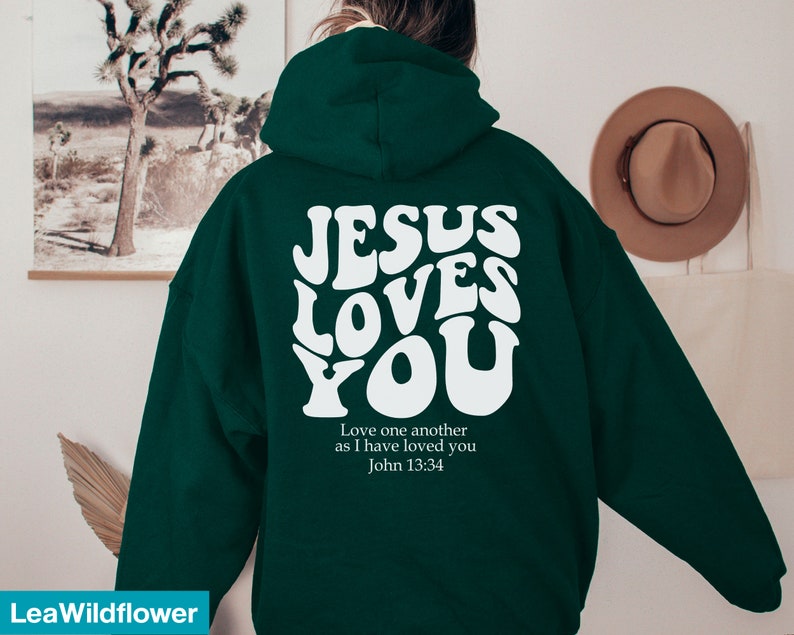 May include: A dark green hoodie with the text "JESUS LOVES YOU" in white, retro-style lettering on the back. Below the text is the quote "Love one another as I have loved you John 13:34".