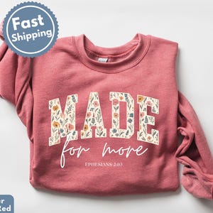 May include: Heather scarlet red sweatshirt with the words "MADE for more" in a floral design. The text "Ephesians 2:10" is below the main text. The sweatshirt has a crew neck and long sleeves.