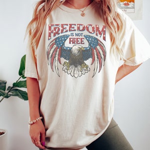 May include: A cream colored t-shirt with a vintage graphic of an eagle with outstretched wings and the text "FREEDOM IS NOT FREE". The eagle is superimposed on a red, white, and blue American flag.