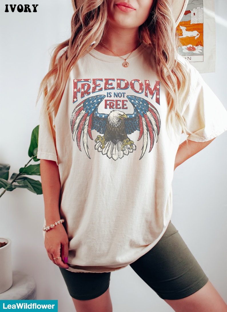 May include: A cream colored t-shirt with a vintage graphic of an eagle with outstretched wings and the text "FREEDOM IS NOT FREE". The eagle is superimposed on a red, white, and blue American flag.