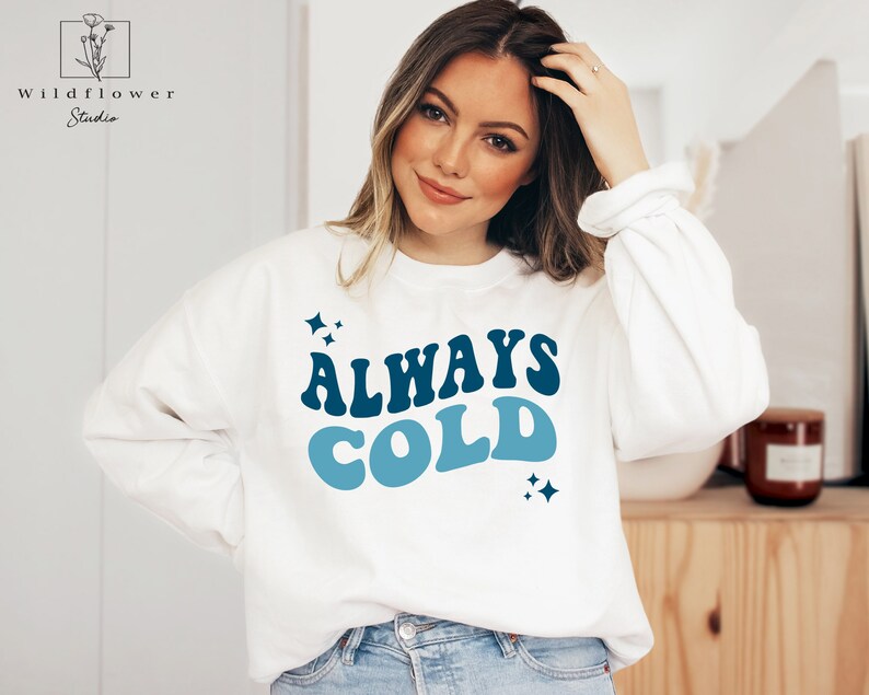 May include: White sweatshirt with blue and dark blue text that reads "ALWAYS COLD" with small blue stars on either side of the text.