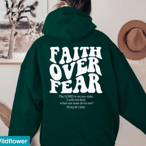 K&ouml;nnte beinhalten: Dunkelgr&uuml;ner Kapuzenpullover mit dem wei&szlig;en Schriftzug "FAITH OVER FEAR" auf der R&uuml;ckseite. Darunter steht ein Bibelvers: "Der HERR ist f&uuml;r mich, ich f&uuml;rchte mich nicht. Was k&ouml;nnen mir Menschen tun? PSALM 118,6."