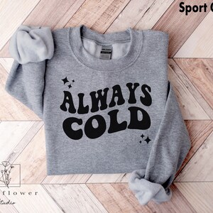 May include: A gray sweatshirt with the text "ALWAYS COLD" in a retro font. The text is in black and the sweatshirt is a sport gray color.