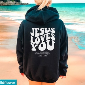 May include: Black hoodie with the text "JESUS LOVES YOU" in white, retro-style lettering. Below the text is the quote "Love one another as I have loved you John 13:34". The hoodie has a hood and long sleeves.