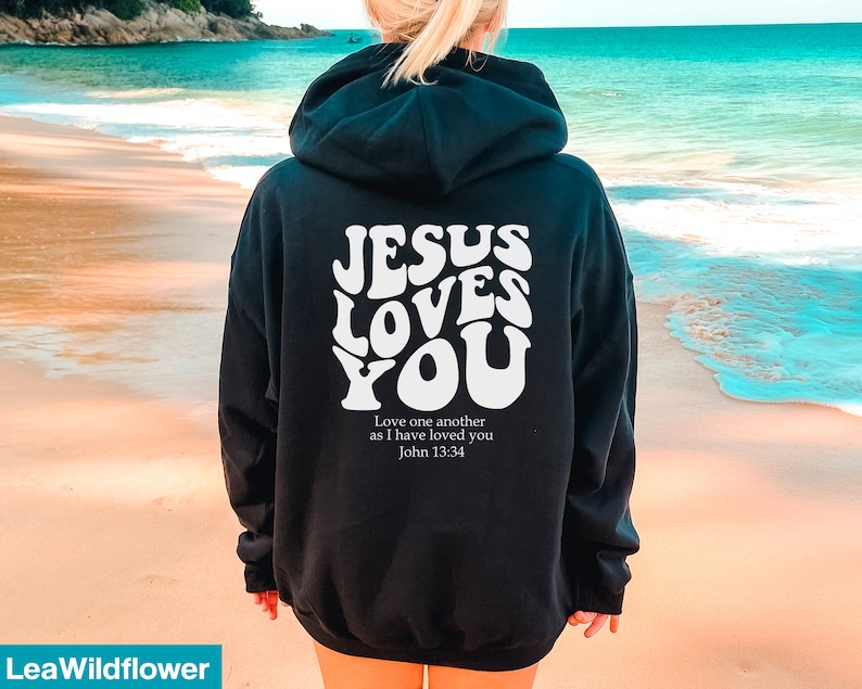 May include: Black hoodie with the text "JESUS LOVES YOU" in white, retro-style lettering. Below the text is the quote "Love one another as I have loved you John 13:34". The hoodie has a hood and long sleeves.