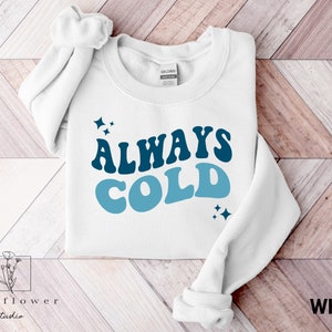 May include: White sweatshirt with blue and white text that reads "ALWAYS COLD" in a retro font. The text is surrounded by small blue stars.