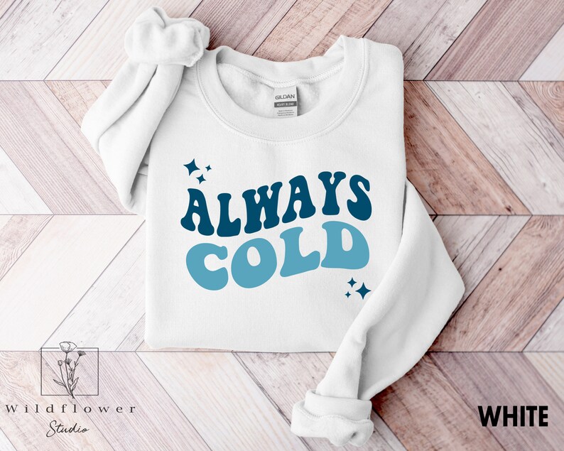 May include: White sweatshirt with blue and white text that reads "ALWAYS COLD" in a retro font. The text is surrounded by small blue stars.