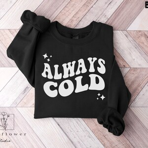 May include: Black sweatshirt with white text that reads "ALWAYS COLD" in a retro font. The text is surrounded by small white stars.