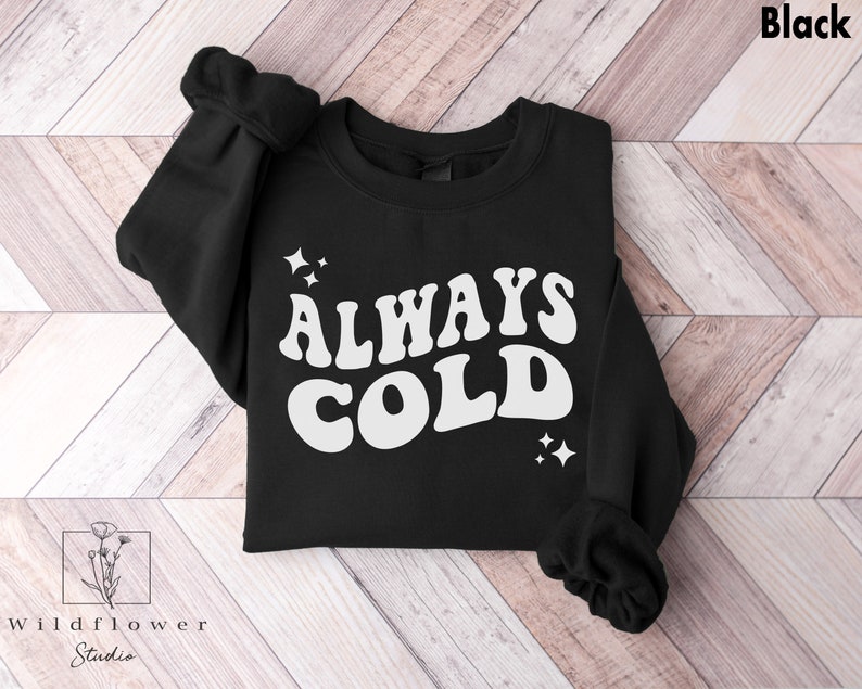 May include: Black sweatshirt with white text that reads "ALWAYS COLD" in a retro font. The text is surrounded by small white stars.
