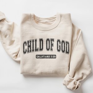 May include: A beige sweatshirt with the text "CHILD OF GOD" in black lettering with a vintage collegiate style font. Below the text is "GALATIANS 3:26" in smaller black lettering.