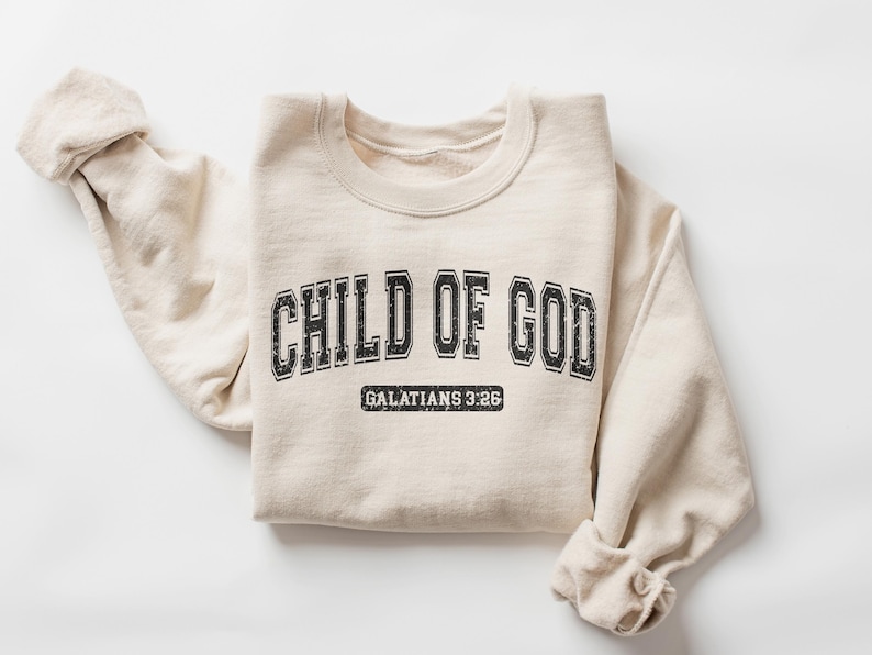 May include: A beige sweatshirt with the text "CHILD OF GOD" in black lettering with a vintage collegiate style font. Below the text is "GALATIANS 3:26" in smaller black lettering.