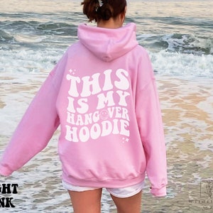 May include: A light pink hoodie with the text "This is my hangover hoodie" in white retro font. The hoodie is worn by a person standing on a beach.