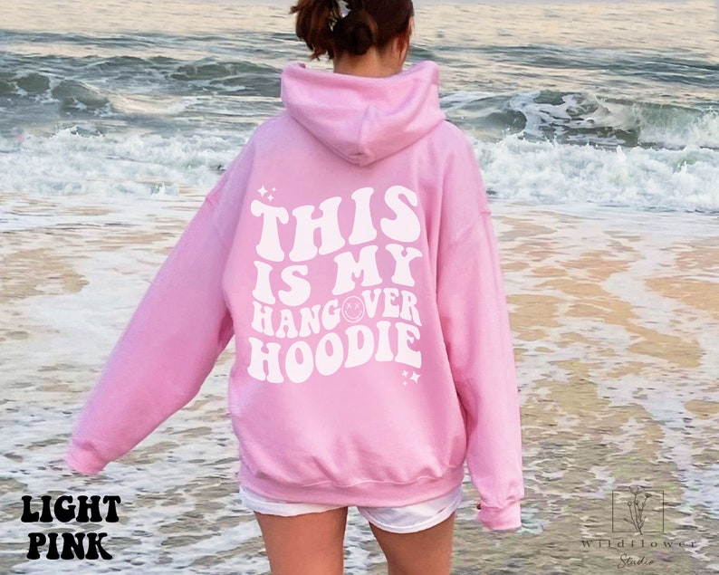 May include: A light pink hoodie with the text "This is my hangover hoodie" in white retro font. The hoodie is worn by a person standing on a beach.