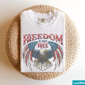May include: White t-shirt with a vintage-style graphic of an eagle with outstretched wings and the text "FREEDOM IS NOT FREE".