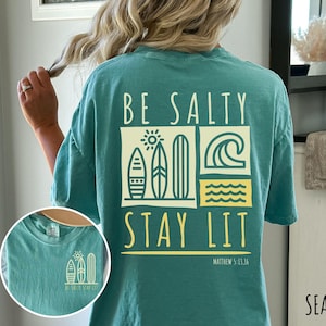 Może przedstawiać: Turkusowa koszulka z nadrukiem graficznym, na kt&oacute;rym widnieje napis "Be Salty Stay Lit" wraz ze słońcem, falami i deskami surfingowymi. Pod grafiką znajduje się tekst "Matthew 5:13-14". Kolor koszulki to "Seafoam".