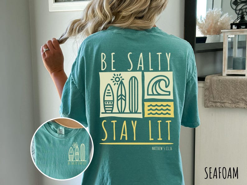Może przedstawiać: Turkusowa koszulka z nadrukiem graficznym, na kt&oacute;rym widnieje napis "Be Salty Stay Lit" wraz ze słońcem, falami i deskami surfingowymi. Pod grafiką znajduje się tekst "Matthew 5:13-14". Kolor koszulki to "Seafoam".