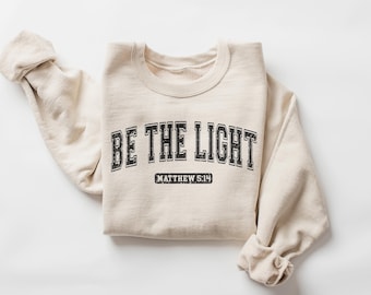 May include: A beige sweatshirt with the text "BE THE LIGHT" in black lettering with a varsity style font. Below the text is "MATTHEW 5:14" in smaller black lettering.