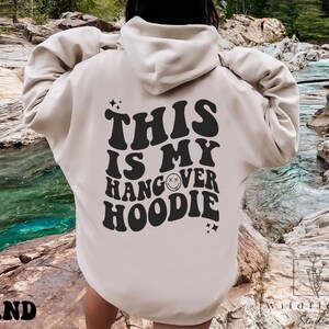 May include: A sand-colored hoodie with the text "This is my hangover hoodie" in black with a smiley face graphic.