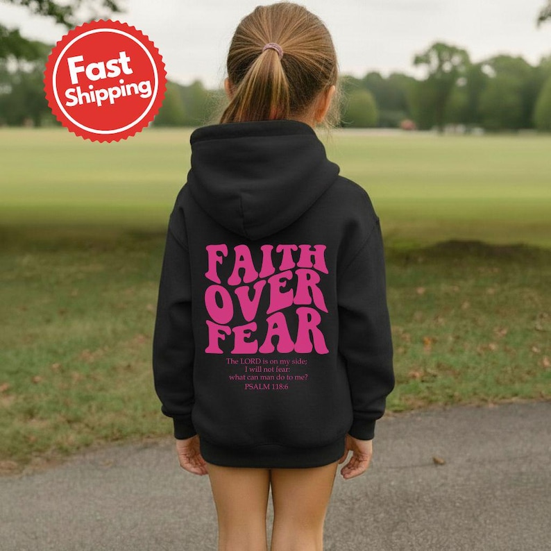 K&ouml;nnte beinhalten: Ein schwarzer Kapuzenpullover mit dem pinkfarbenen Schriftzug "FAITH OVER FEAR" und einem Bibelvers. Die Person tr&auml;gt den Pullover in einer Gr&uuml;nfl&auml;che. Der Pullover hat eine Kapuze und lange &Auml;rmel.