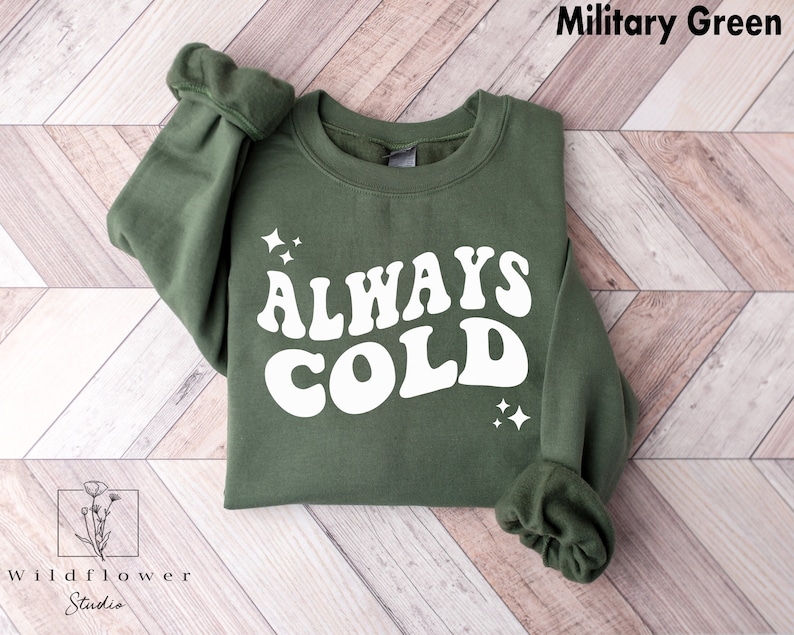 May include: A green sweatshirt with the text "ALWAYS COLD" in white retro-style lettering. The sweatshirt has a crew neck and long sleeves.
