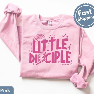 May include: Light pink sweatshirt with the words "LITTLE DISCIPLE" in bright pink. The design includes a rainbow, cross, crown, and lightning bolt. The sweatshirt has long sleeves and a crew neck.